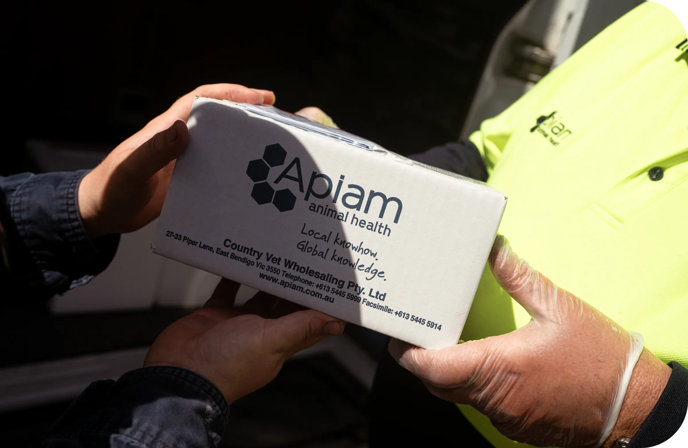 A box of Apiam Animal Health veterinary supplies being handed from one person to another.